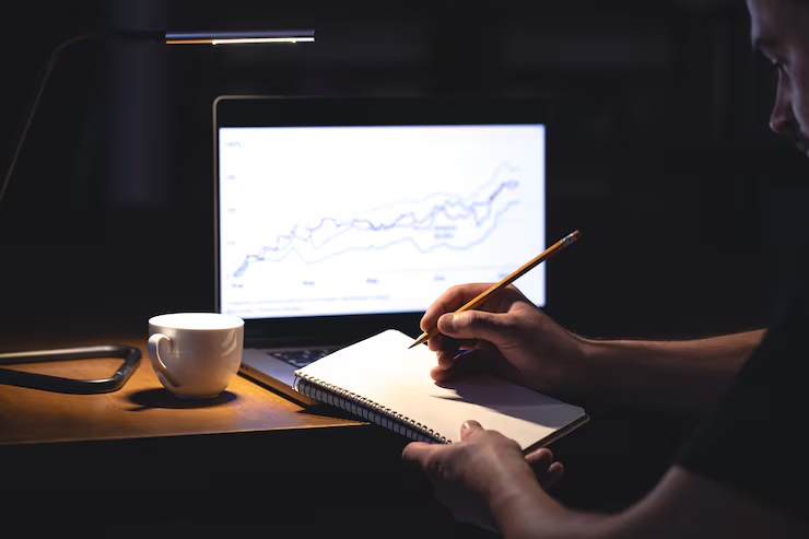 A trader takes notes in front of a laptop showing a rising chart, late at night